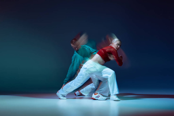Portrait of young man and woman dancing hip-hop isolated over dark blue background with mixed lights. Training class. Concept of movement, youth culture, active lifestyle, action, street dance, ad - Photo, Image