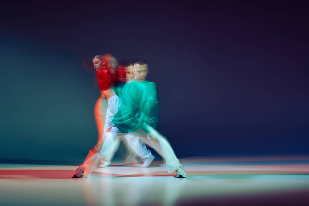 Portrait of young man and woman dancing isolated over dark blue background with mixed lights. Cooperation. Concept of movement, youth culture, active lifestyle, action, street dance, ad - Photo, image