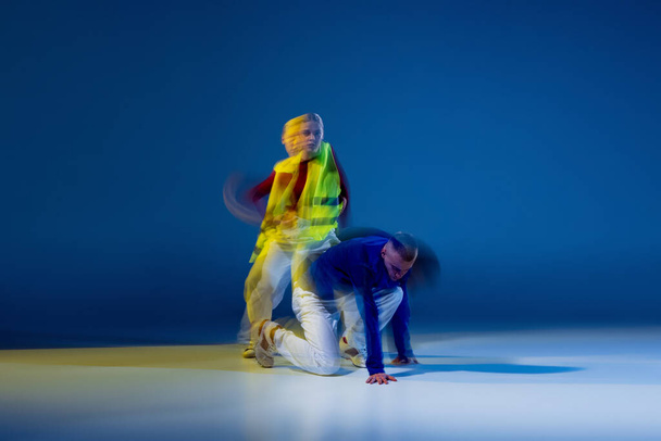 Portrait of young couple, man and woman dancing hip-hop isolated over dark blue background in yellow neon with mixed lights. Concept of movement, youth culture, active lifestyle, action, street dance - Photo, Image