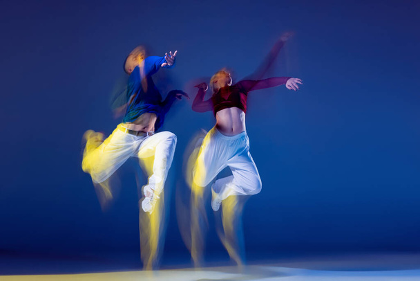 Retrato de jovem e mulher dançando isolado sobre fundo azul escuro com luzes mistas. Num salto. Conceito de movimento, cultura juvenil, estilo de vida ativo, ação, dança de rua, anúncio - Foto, Imagem
