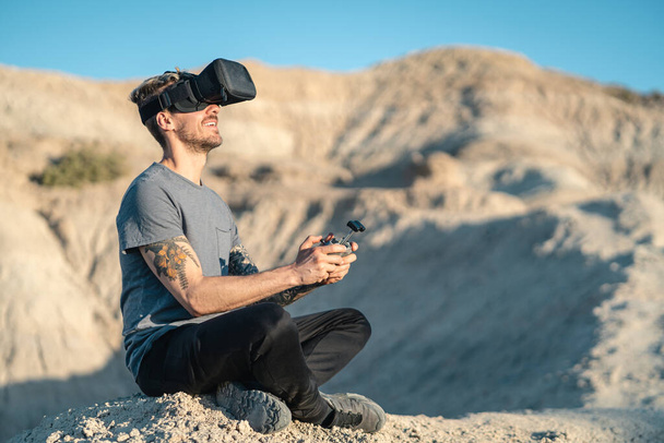 Happy young adult man flying drone with VR mask while sitting on outdoors badlands area. Copy Space on right side - Фото, зображення