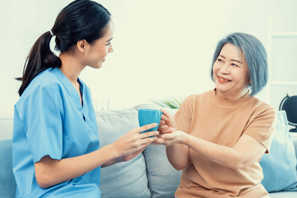Female care taker serving her contented senior patient with a cup of coffee at home, smiling to each other. Medical care for pensioners, Home health care service. - Foto, imagen