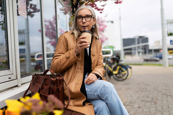 Kahve içen yaşlı bir kadın mağazanın yanındaki bankta oturuyor.. - Fotoğraf, Görsel