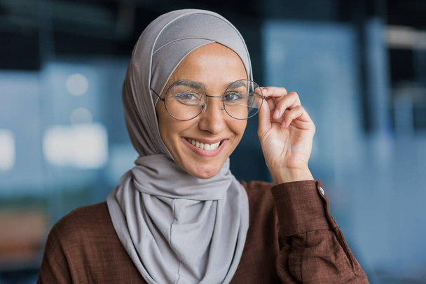 Close-up photo of a young beautiful woman in a hijab and glasses looking at the camera and smiling, a student close-up studying inside a modern university. - Photo, Image
