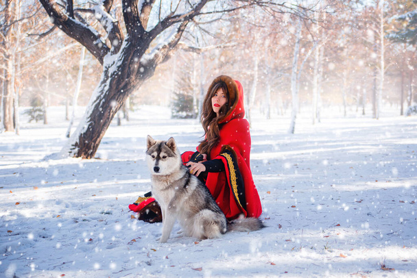 A young woman in a red hooded raincoat with a gray husky dog on the background of a winter forest and snowfall. - Fotó, kép