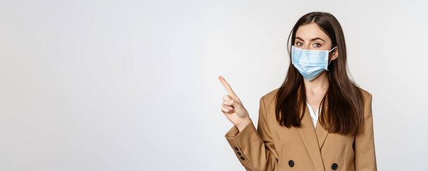 Ο ιός του κερατοειδούς και οι άνθρωποι το αντιλαμβάνονται. Πορτρέτο της γυναίκας των επιχειρήσεων στο χώρο εργασίας φορώντας μάσκα προσώπου, δείχνοντας το δάχτυλο αριστερά στο λογότυπο, banner της εταιρείας, λευκό φόντο. - Φωτογραφία, εικόνα