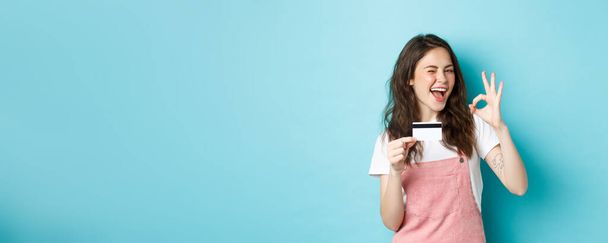 Fröhliches brünettes Mädchen zeigt Plastikkreditkarte und Okay-Zeichen, zwinkert und lächelt selbstbewusst, empfiehlt Logo, steht über blauem Hintergrund. - Foto, Bild