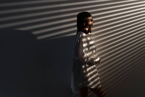 Πορτρέτο μιας σαγηνευτικής γυναίκας που φοράει ανδρικό πουκάμισο ποζάροντας πάνω από γκρι φόντο, σκιά από περσίδες, σέξι πρωινό βλέμμα. Αντιγραφή χώρου - Φωτογραφία, εικόνα