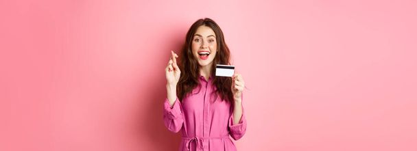 Image d'une jeune femme pleine d'espoir montrant une carte de crédit en plastique et des doigts croisés pour la bonne chance, faisant vœu et souriant, debout sur fond rose. Espace de copie - Photo, image