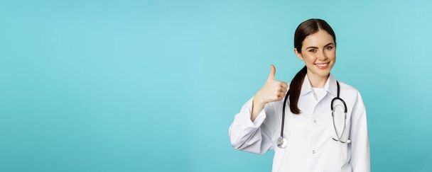 Усміхнена дівчина-лікар, працівник охорони здоров'я зі стетоскопом, показує великий палець у схваленні, рекомендує клініку або аптеку, стоячи на бірюзовому фоні
. - Фото, зображення