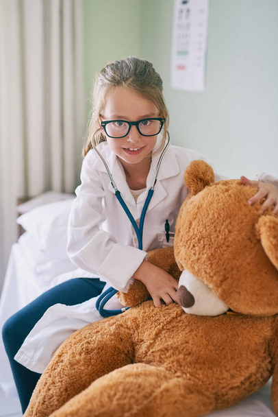 Μαθαίνω ιατρική από μικρή ηλικία. Ένα κοριτσάκι που παριστάνει τον γιατρό ενώ εξετάζει το αρκουδάκι της. - Φωτογραφία, εικόνα