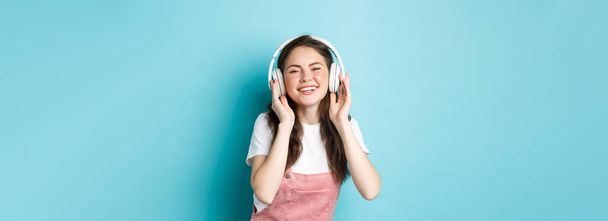 Aranyos kaukázusi lány tavaszi ruhában, zenét hallgat fejhallgatóban, mosolyog elégedett a kamerával, áll a kék háttér felett. - Fotó, kép
