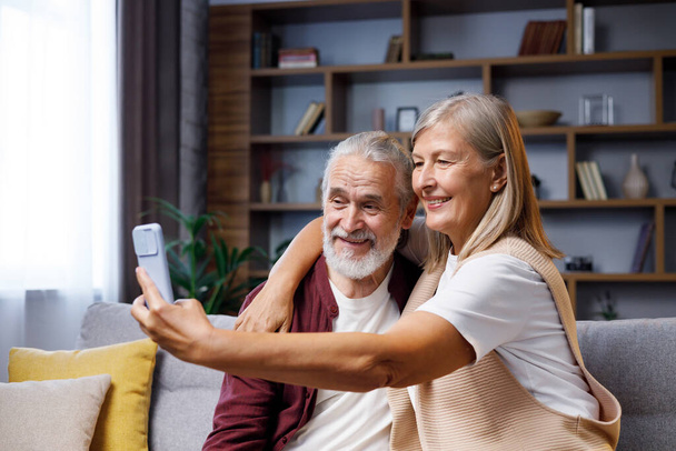 Mature happy couple fooling around on smartphone camera. Gray-haired husband and wife are talking making funny selfies. Video call on smartphone. Happy pensioners - 写真・画像