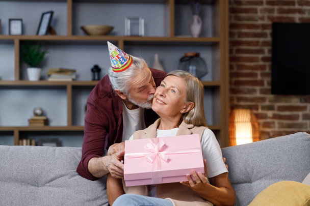 Caring old husband giving romantic birthday present to excited mature wife, loving older man surprise happy middle aged woman, open pink box, kiss, hug husband, celebrating together - Valokuva, kuva