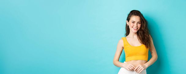 Mulher elegante jovem pronto para férias de verão, vestindo top amarelo e sorrindo alegre para a câmera, de pé contra o fundo azul. - Foto, Imagem