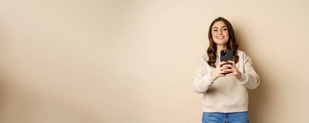 Happy smiling girl recording video, taking picture on smartphone and looking at camera, standing over beige background. Copy space - 写真・画像