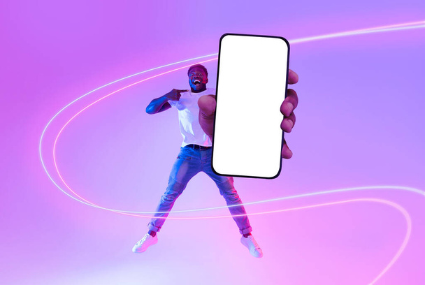 Joyeux jeune homme afro-américain en tenue décontractée sautant et montrant tout nouveau smartphone avec écran blanc vierge pour la publicité, posant au néon, collage, maquette, photo pleine longueur - Photo, image