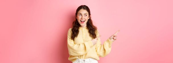 Happy good-looking girl demonstrate information, pointing fingers left and staring at promotional text with pleased smile, standing against pink background. - Photo, Image
