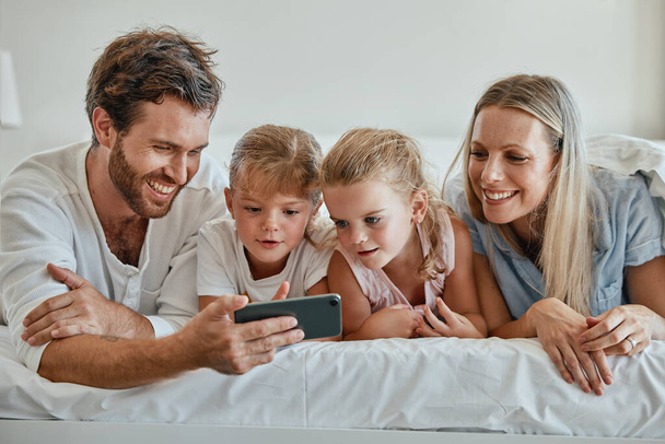 Szczęśliwa rodzina z dziećmi, telefon i leżenie w łóżku w domu, matka, ojciec i dziewczynka dzieci oglądając razem film. Miłość, zabawa i rodzina z tatą, mama i córki na telefon wideo smartfona w sypialni - Zdjęcie, obraz