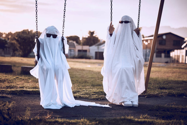 Park, halloween and friends in a ghost costume in a kids playground in celebration of a scary fantasy holiday. Glasses, creepy and crazy couple in horror, creative and spooky white sheets on a swing. - Foto, Imagem