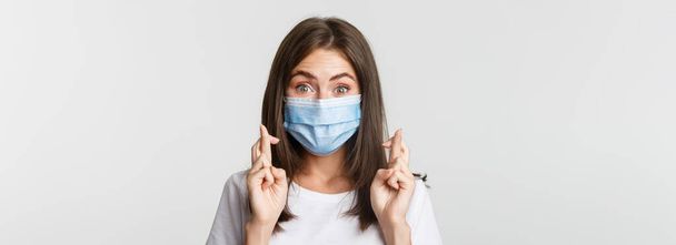 Covid-19, health and social distancing concept. Close-up of hopeful smiling girl in medical mask, cross fingers good luck. - Фото, изображение
