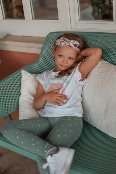 Bela criança menina na moda em roupas de moda com uma camiseta branca, leggings e tênis brancos senta-se e descansar em um banco verde com travesseiros perto da casa - Foto, Imagem