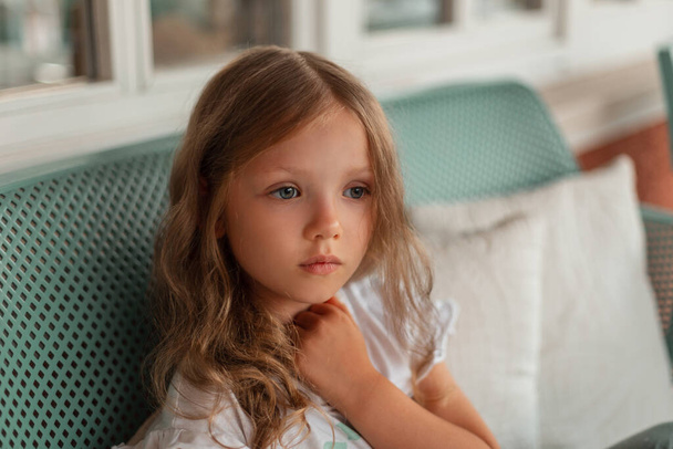Hämmästyttävä lapsi muotokuva kaunis pieni tyttö sinisilmäinen istuu penkillä talon ulkopuolella. Tunteet - Valokuva, kuva