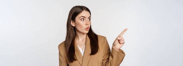 Portrait of businesswoman pointing finger left, showing corporate banner, logo, standing in brown suit over white background. - 写真・画像