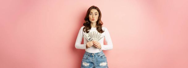 Кредити та мікрокредитування. Усміхнена красива дівчина показує гроші, готівку в руках і виглядає захоплено, стоїть над рожевим тлом
. - Фото, зображення
