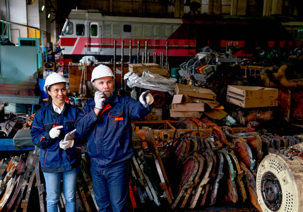 Caucasian factory worker or technician use walkie talkie and stand beside Asian factory woman in front of the train and spare part of machine in workplace area. - Photo, Image
