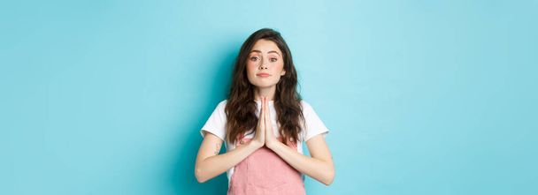 Please help me. Cute brunette woman asking for favour, holding hands in pray, begging you to buy something, standing against blue background. - Photo, Image
