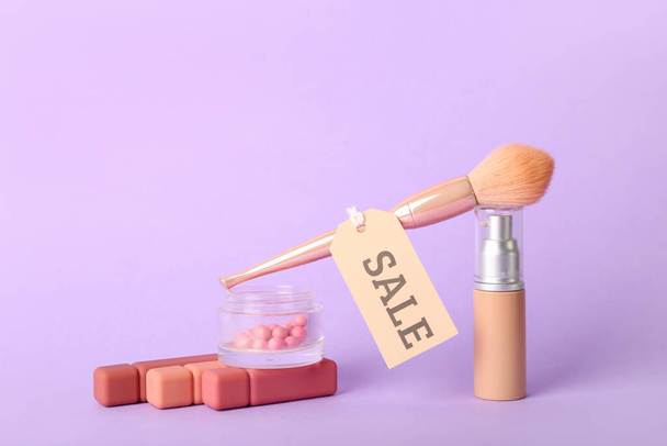 Dekorative Kosmetik mit Make-up-Pinsel und Verkaufsetikett auf fliederfarbenem Hintergrund - Foto, Bild