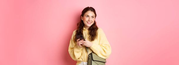 Stylowa, nowoczesna dziewczyna z uśmiechem na twarzy, romantyczna, czatująca na smartfonie z aplikacją randkową, stojąca z torebką na różowym tle. - Zdjęcie, obraz