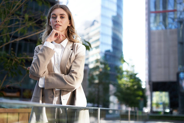 Silná a sebevědomá podnikatelka hledící na kameru, držící se za ruku, stojící před kancelářskou budovou v béžovém obleku. - Fotografie, Obrázek