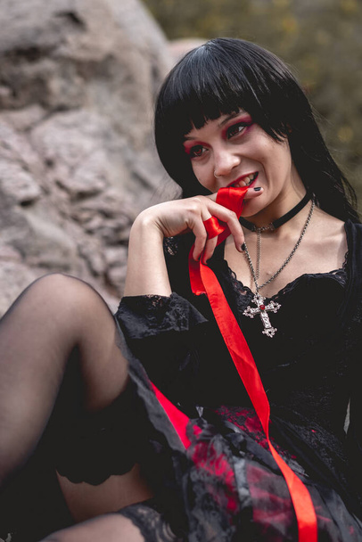 Young, sexy and vampiric hispanic goth girl with black dress seated and playing with "blood" (red ribbon) in a wood bench between big rocks under sunlight - Photo, Image