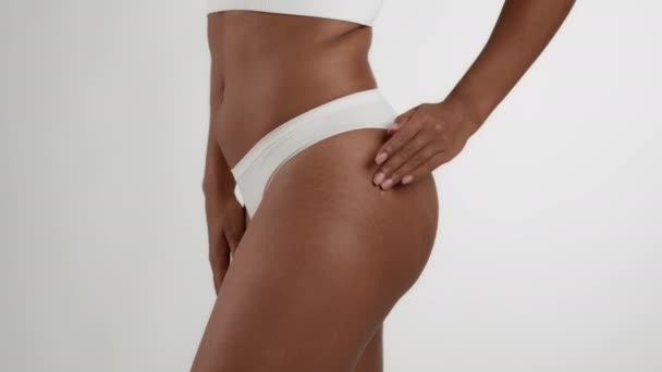 Antycellulitowa pielęgnacja ciała. Zbliżenie strzał nierozpoznawalny Afroamerykanka kobieta w bieliźnie tarcie skóry podnoszenie krem na biodrach skóry, białe tło studio, zwolnione tempo - Materiał filmowy, wideo