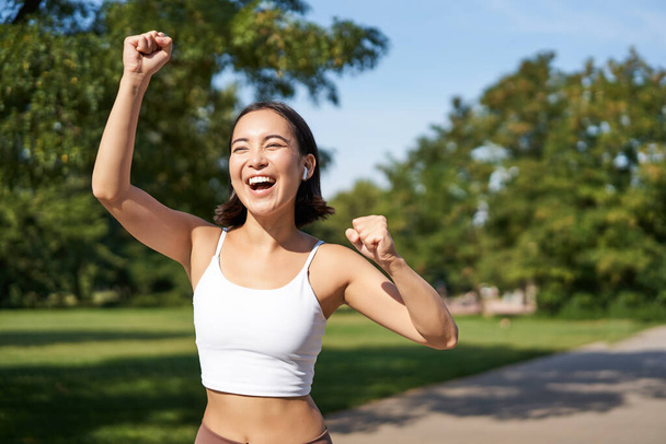 Happy fitness girl achieve goal, finish marathon, running with hands up, celebrating victory while jogging, triumphing in park. - Foto, immagini