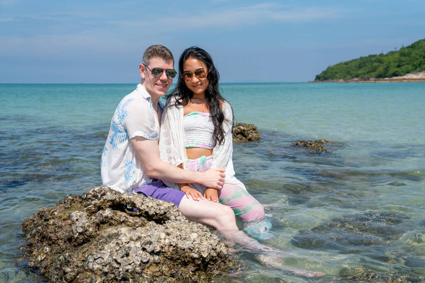 Caucasian man sit on the rock with Asian woman and look at camera with smiling on day time during honey moon vacation or holiday. - Φωτογραφία, εικόνα