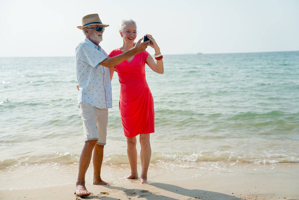 Homem sênior segurar telefone celular e ação olhar como photoshoot selfie com a mulher sênior na praia durante o relaxamento em férias ou férias com felicidade. - Foto, Imagem