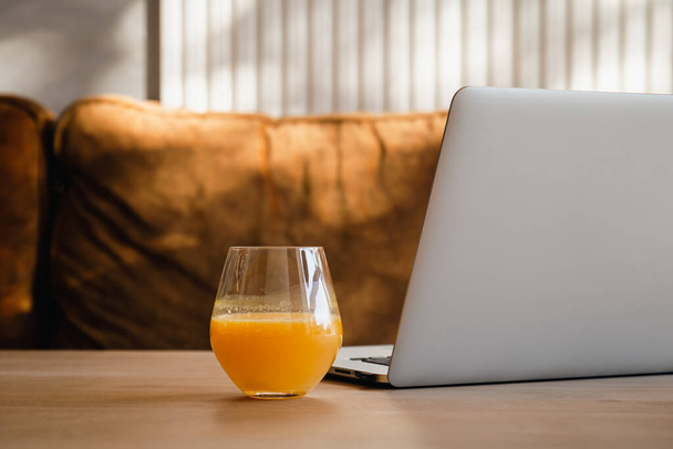 Szklanka soku pomarańczowego w pobliżu laptopa na stole, we wnętrzu ze światłem słonecznym, koncepcja pracy rano. - Zdjęcie, obraz