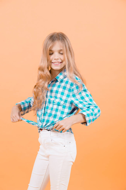 Girl model with closed eyes, blond long hair smile in blue plaid shirt and white pants on orange background. Kid fashion, style, trend concept. Beauty, look, hairstyle. - Fotoğraf, Görsel