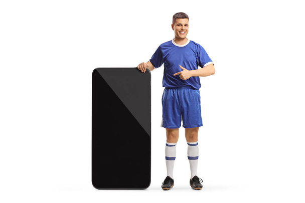Man in a football jersey standing next to a big smartphone and pointing at the screen isolated on white background - Фото, зображення