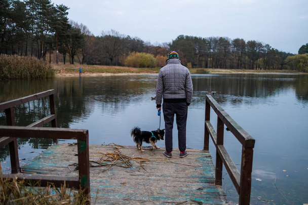 El dueño del perro y su amigo perro están de pie en el muelle de madera cerca del lago y disfrutando del paisaje durante su paseo en el tiempo de temporada de otoño. Imagen del concepto humano y animal doméstico. - Foto, Imagen