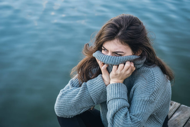 An attractive young woman in a sweater sits by the river against the background of the water, the cold season. - Foto, imagen