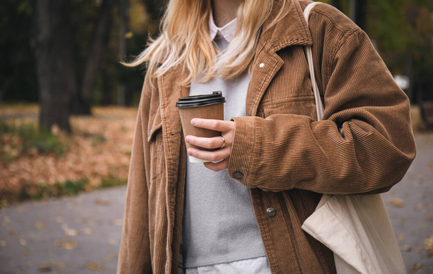 Genç bir kadın, parkta yürürken elinde tek kullanımlık bir bardak kahve tutuyor.. - Fotoğraf, Görsel