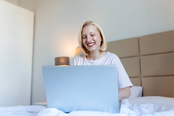 Atrakcyjna kobieta w białej koszuli używająca laptopa i uśmiechnięta rano. Czat wideo i freelance pracy z domu koncepcji pięknej młodej kobiety z laptopem w łóżku - Zdjęcie, obraz