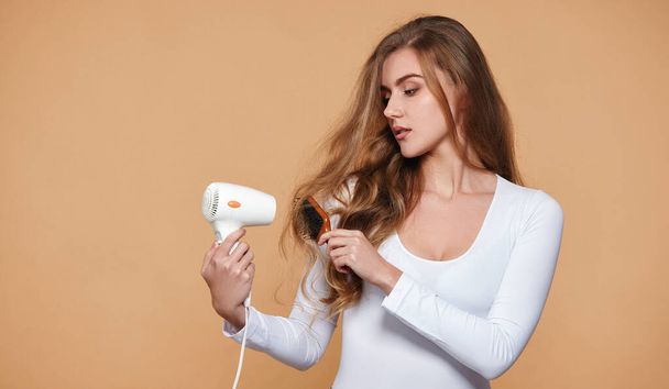 lovely woman with strong healthy curly hair using white hair dryer on beige background - Foto, Imagem