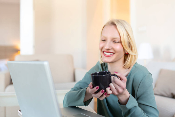 Pozytywna młoda kobieta freelancer oglądanie wideo w blogu na netbook i o kawę w komfortowym domu. Piękna młoda kobieta biznesu pracująca na laptopie.Freelancer podłączony do Internetu - Zdjęcie, obraz
