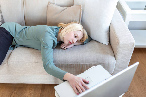 Wyczerpana młoda kobieta leżąca na sofie, korzystająca z laptopa, zbyt zmęczona lub znudzona pracą online w domu, wolną przestrzenią. Pracoholizm, chroniczne zmęczenie, przepracowanie koncepcji pracy na odległość - Zdjęcie, obraz