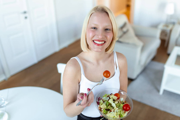 Портрет счастливой игривой девушки, которая ест свежий салат из миски на кухне. Красивая подтянутая женщина ест здоровый салат после тренировки - Фото, изображение